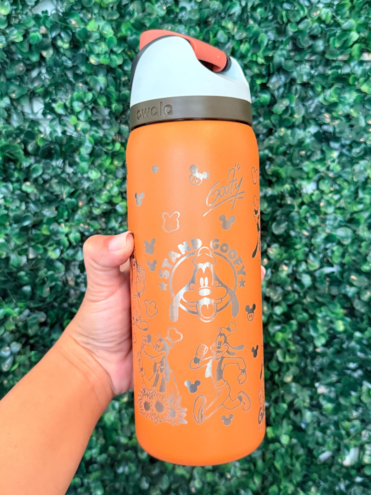 Orange Goofy-inspired engraved Owala in front of a green hedge