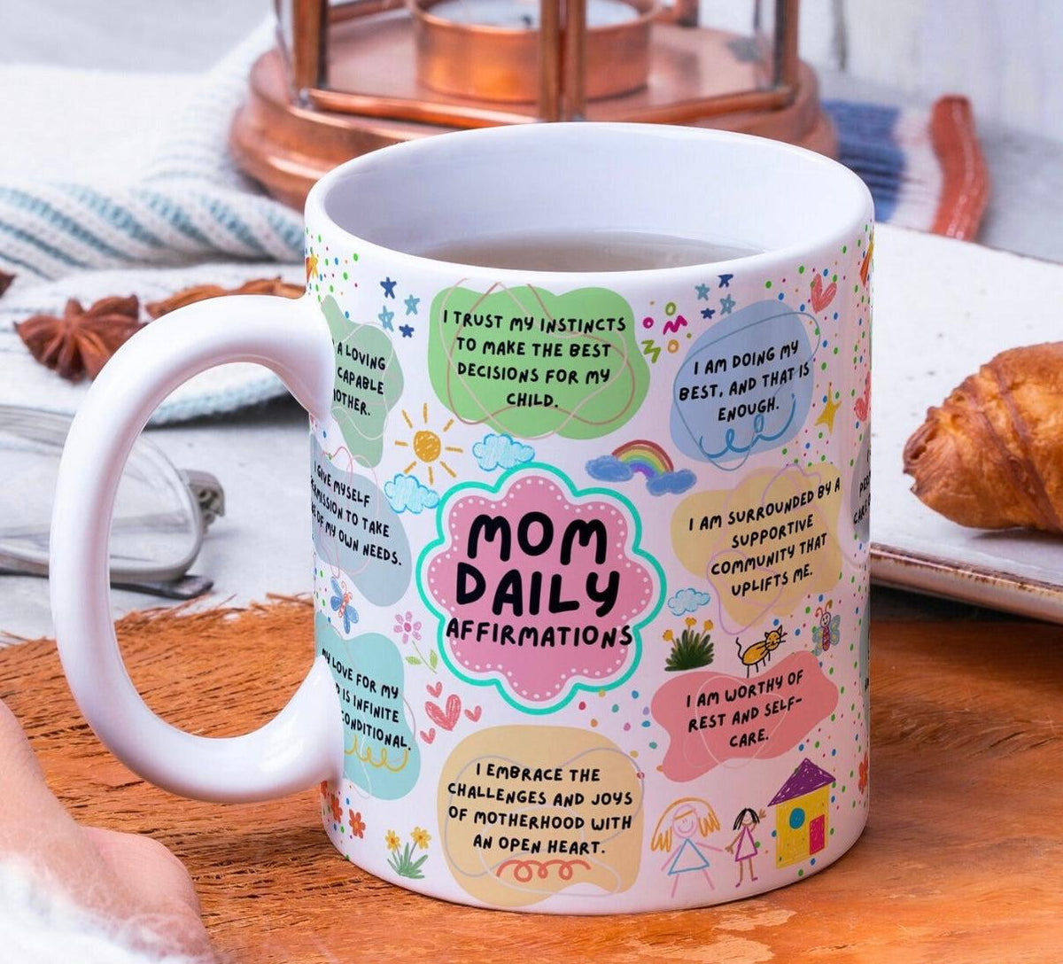 White ceramic mug with colorful Mom Daily Affirmations print next to tea