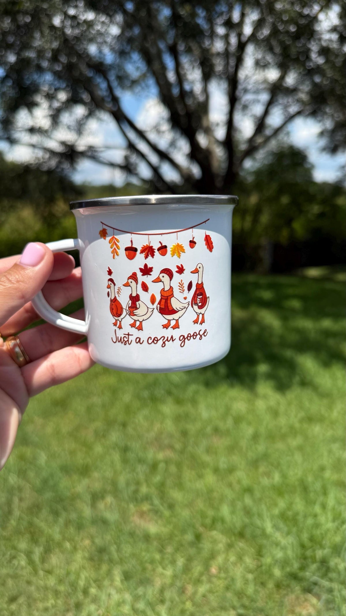 White enamel camp mug with a 'Just a cozy goose' fall design, held outdoors