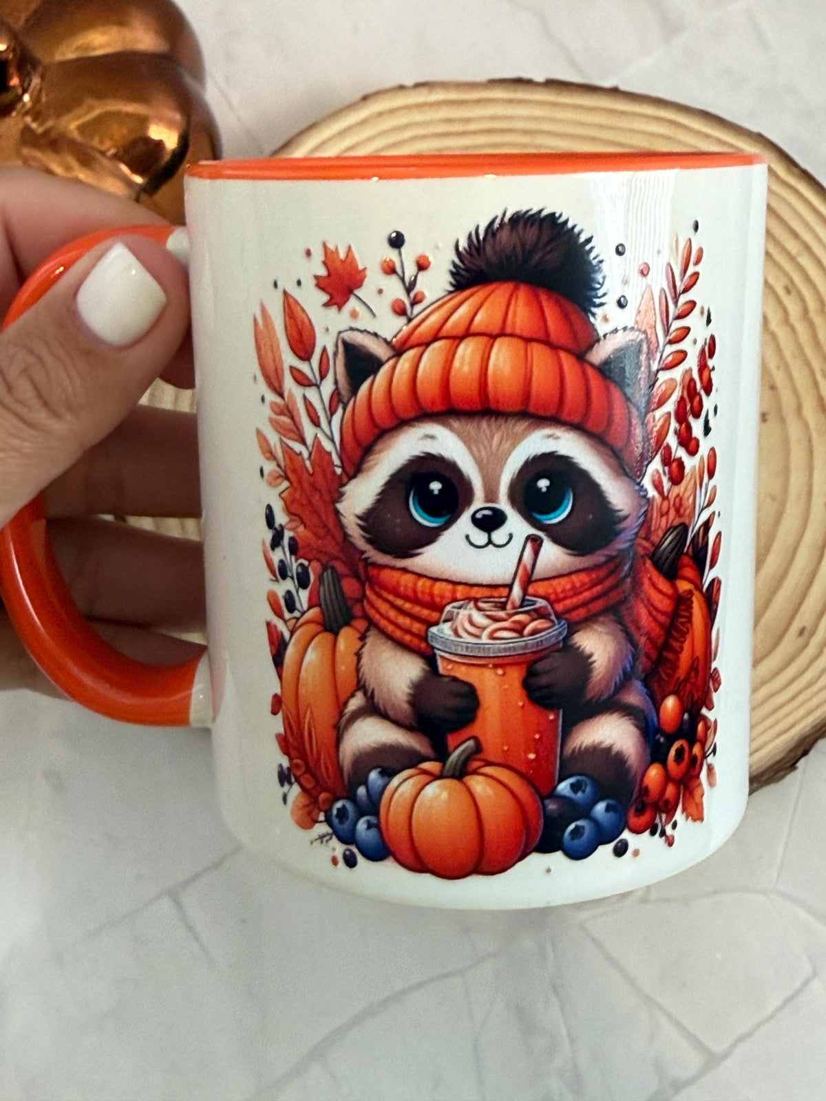 White ceramic mug with orange handle and an autumn raccoon in a knit hat